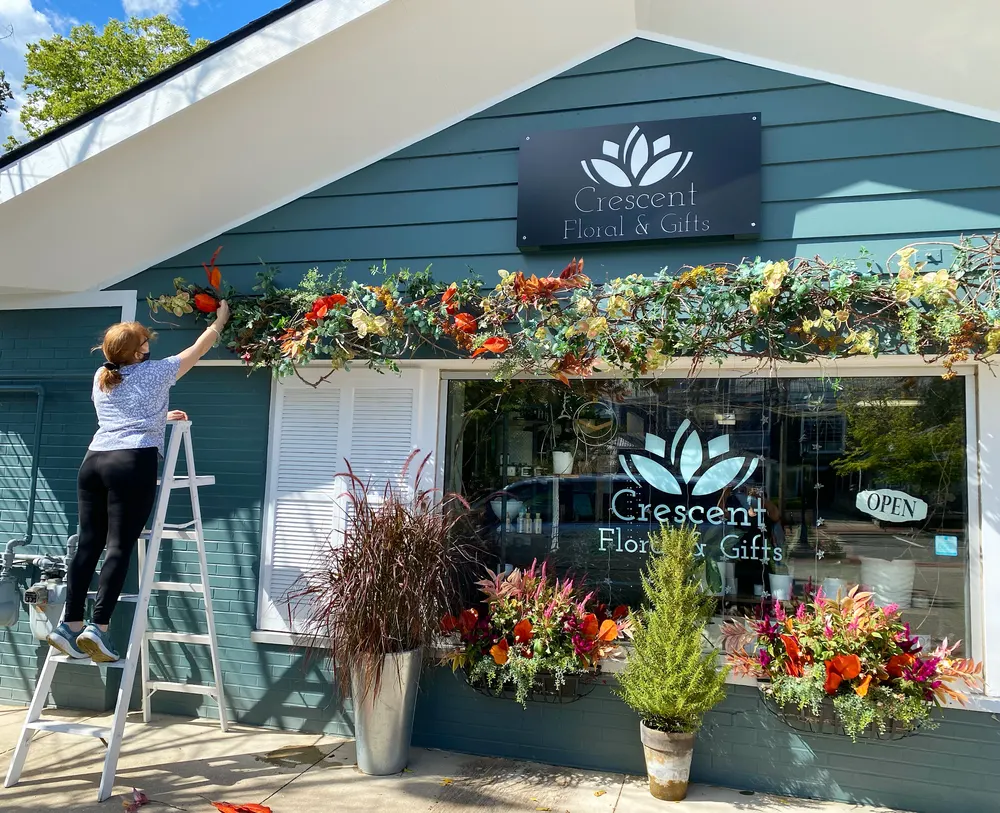 Crescent Floral & Gifts storefront — an employee on a white ladder hangs an autumn garland over the display window of the deep-teal building, vivid planters below the lotus sign