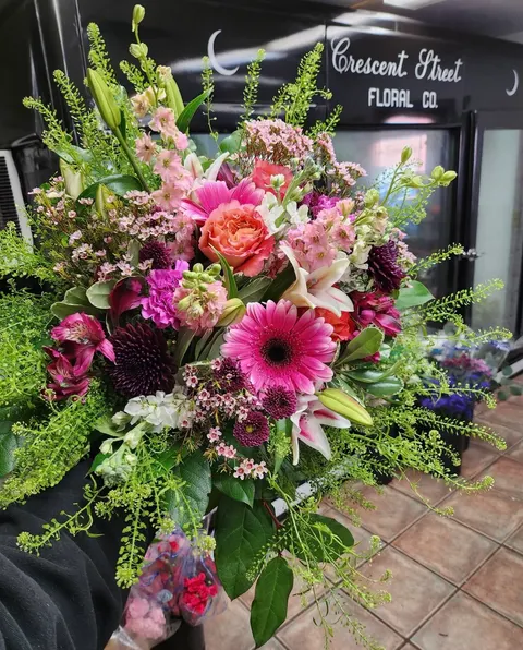 A vivid hand-held bouquet of magenta gerberas, lilies and ranunculus in front of the black Crescent Street Floral Co. cooler sign