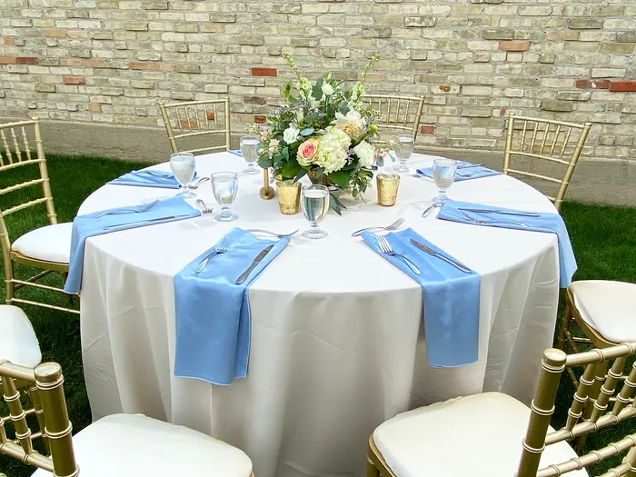 Outdoor wedding table with white linens, periwinkle napkins, gold chiavari chairs and a blush centerpiece against a cream brick wall